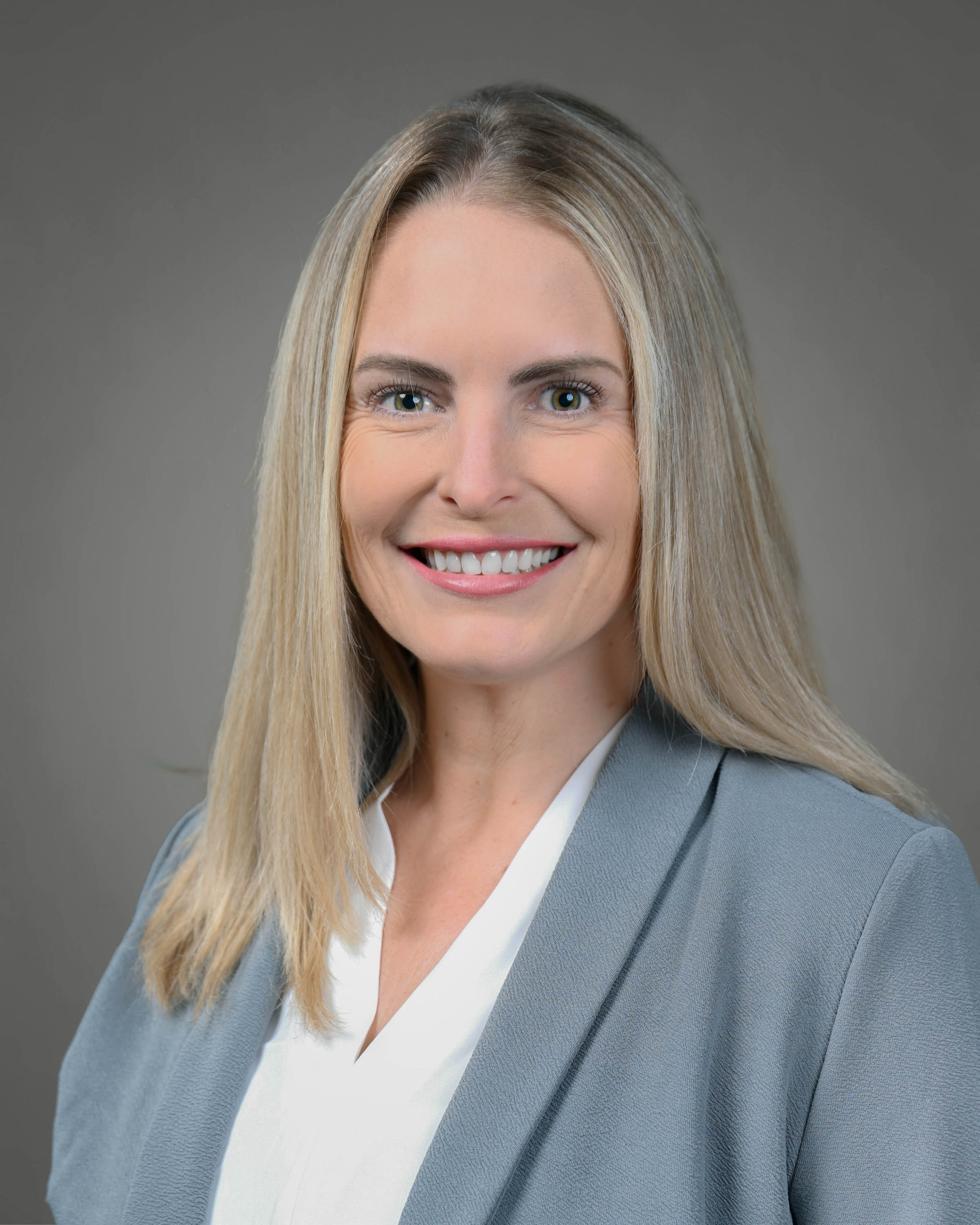 Professional headshot of a smiling woman with long blonde hair, wearing a light gray blazer and white blouse against a gray studio background.